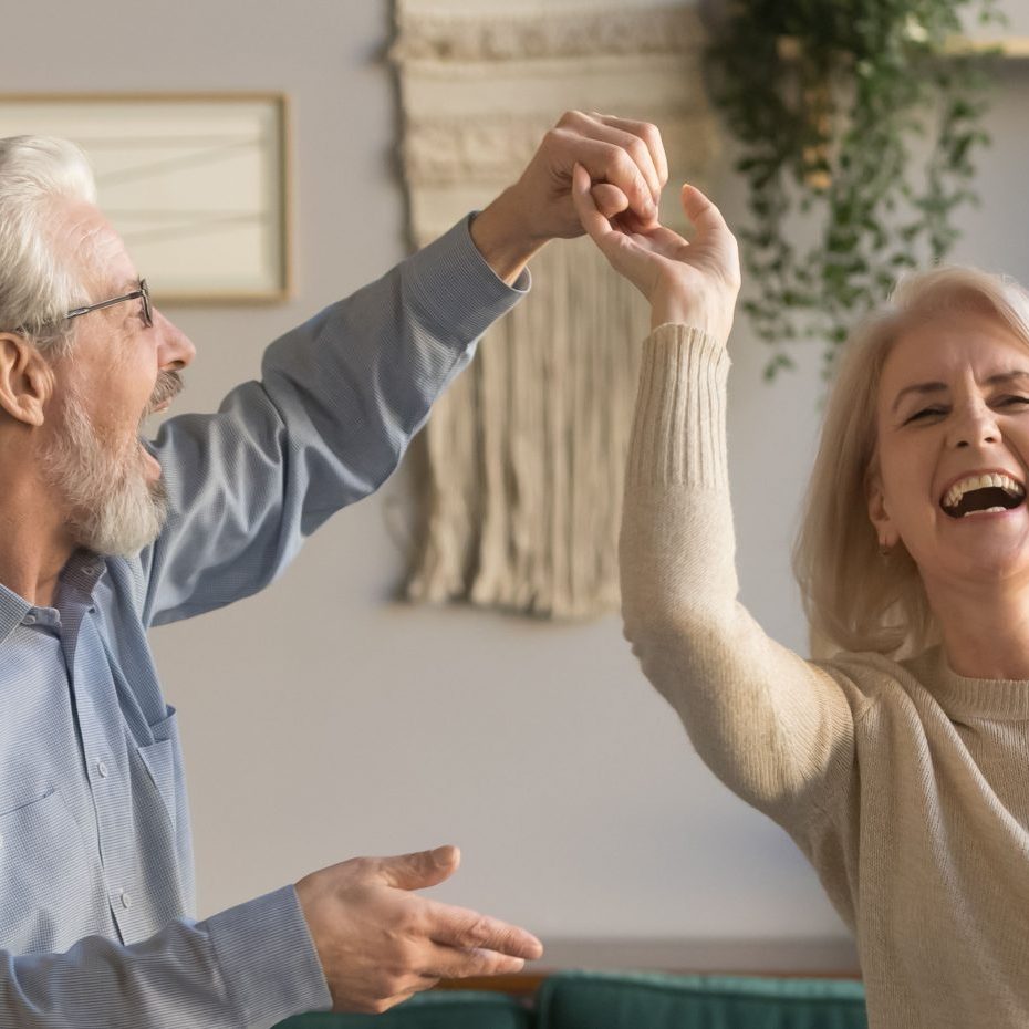 Joyful active old retired romantic couple dancing laughing in living room, happy middle aged wife and elder husband having fun at home, smiling senior family grandparents relaxing bonding together